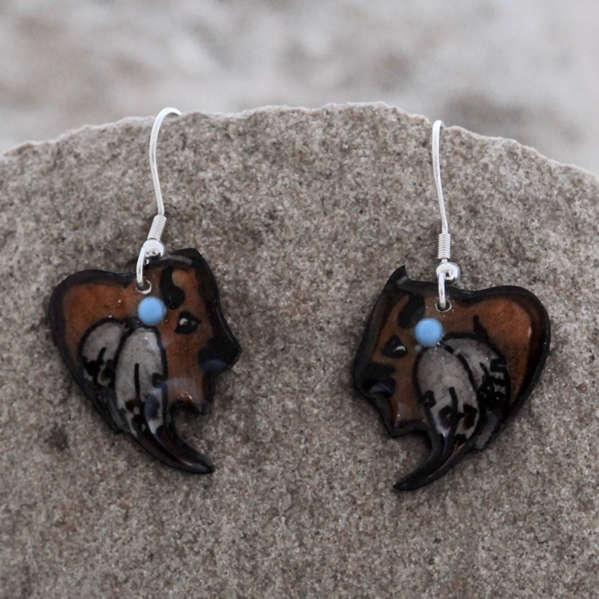 A pair of handmade glass dangle earrings shaped like stylized dog profiles, resting on a textured gray stone. The earrings feature brown and black glass with white and gray marbled details, a small light blue bead, and silver fishhook wires.