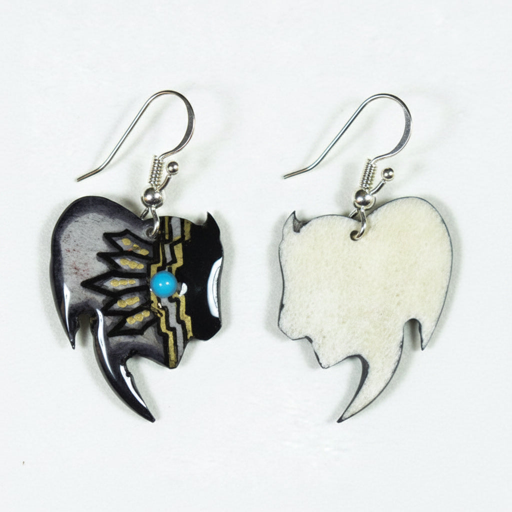 A pair of Native American style dangle earrings on a white background. One earring features a black and gray shell with gold inlay and a small turquoise bead, while the other is a white shell with black inlay.